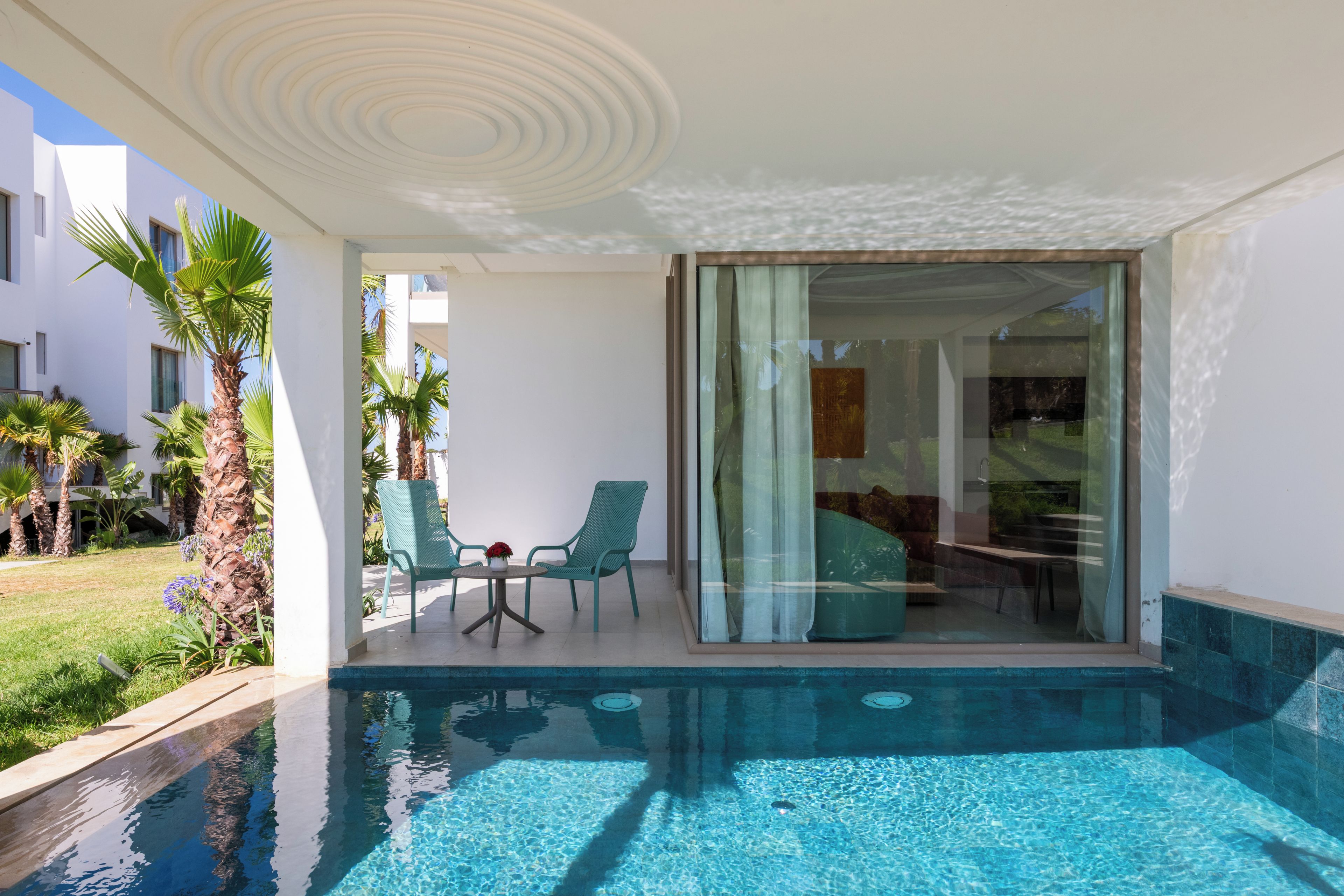 Un área de piscina moderna con una puerta de vidrio que da a una habitación, rodeada de palmeras y una piscina azul cristalina.