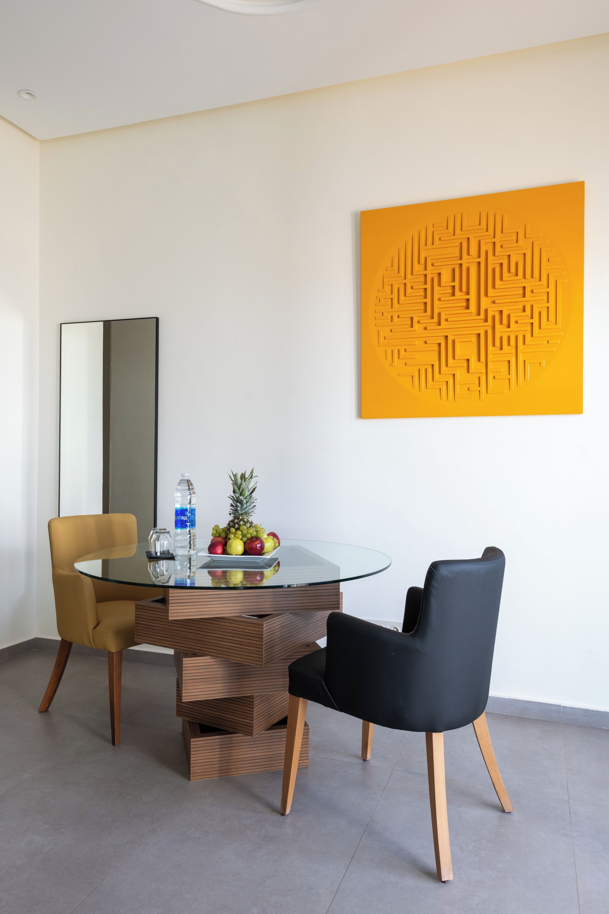 A modern dining area with a glass table, two chairs, a fruit bowl, a water bottle, and a decorative wall piece.