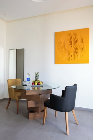A modern dining area with a glass table, two chairs, a fruit bowl, a water bottle, and a decorative wall piece. - Le Tanger By Mnar Palm (Al Bahraoyine)