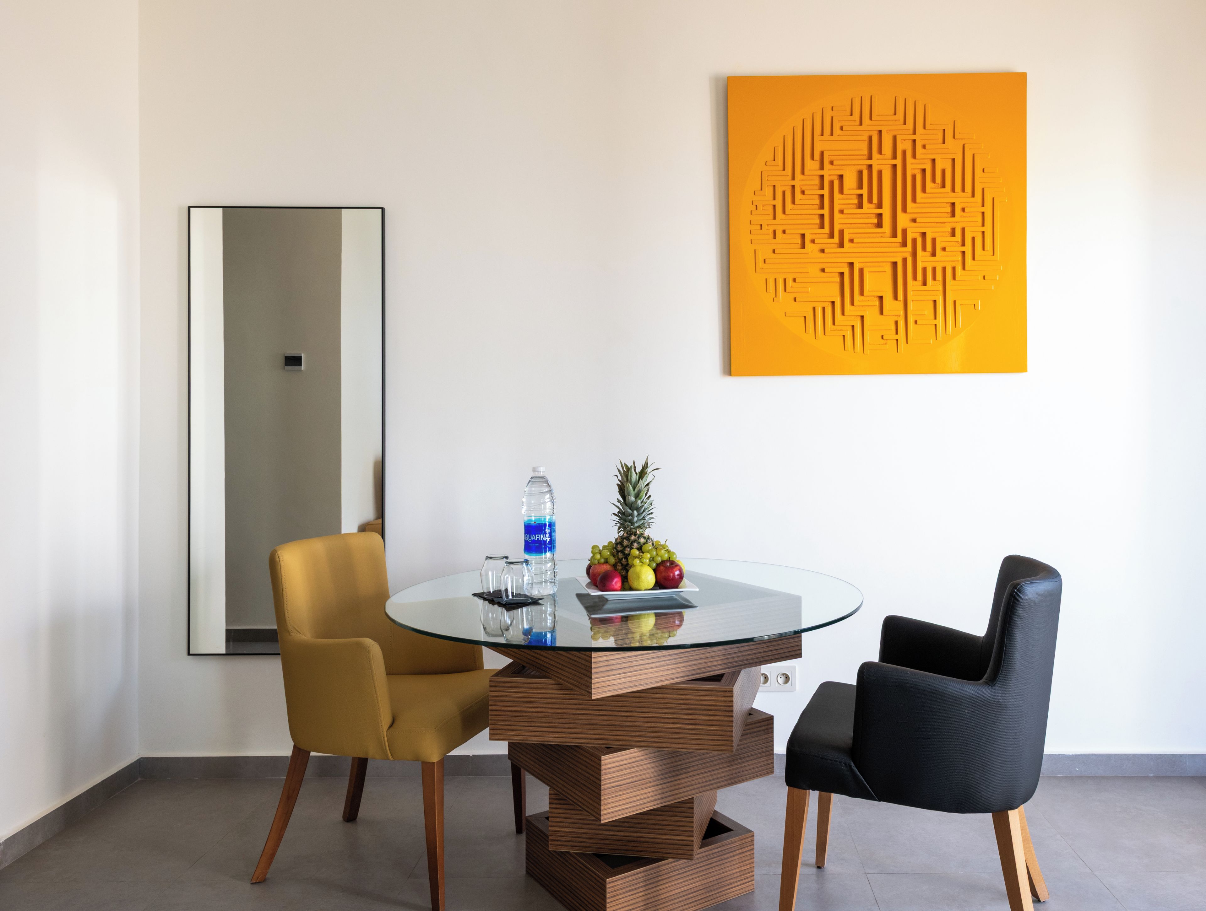 Un comedor moderno con una mesa de vidrio, dos sillas, un cuenco de frutas y una botella de agua.
