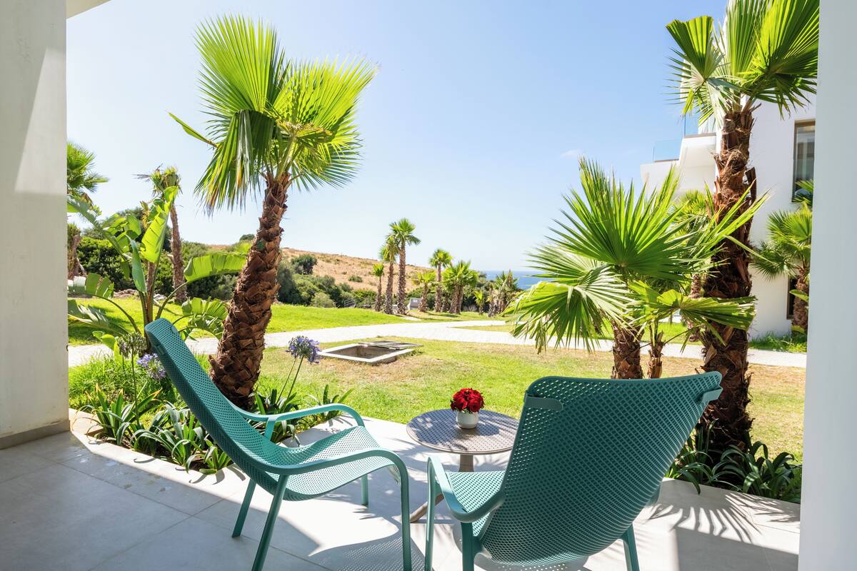 A patio with two green chairs, a small table with a vase of red flowers, and a view of a garden with palm trees and a white building.