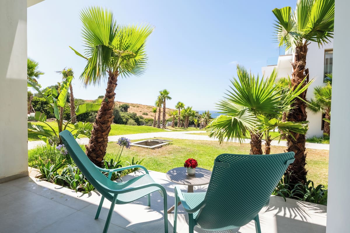 A patio with two green chairs and a table, surrounded by palm trees and a view of a landscaped garden.