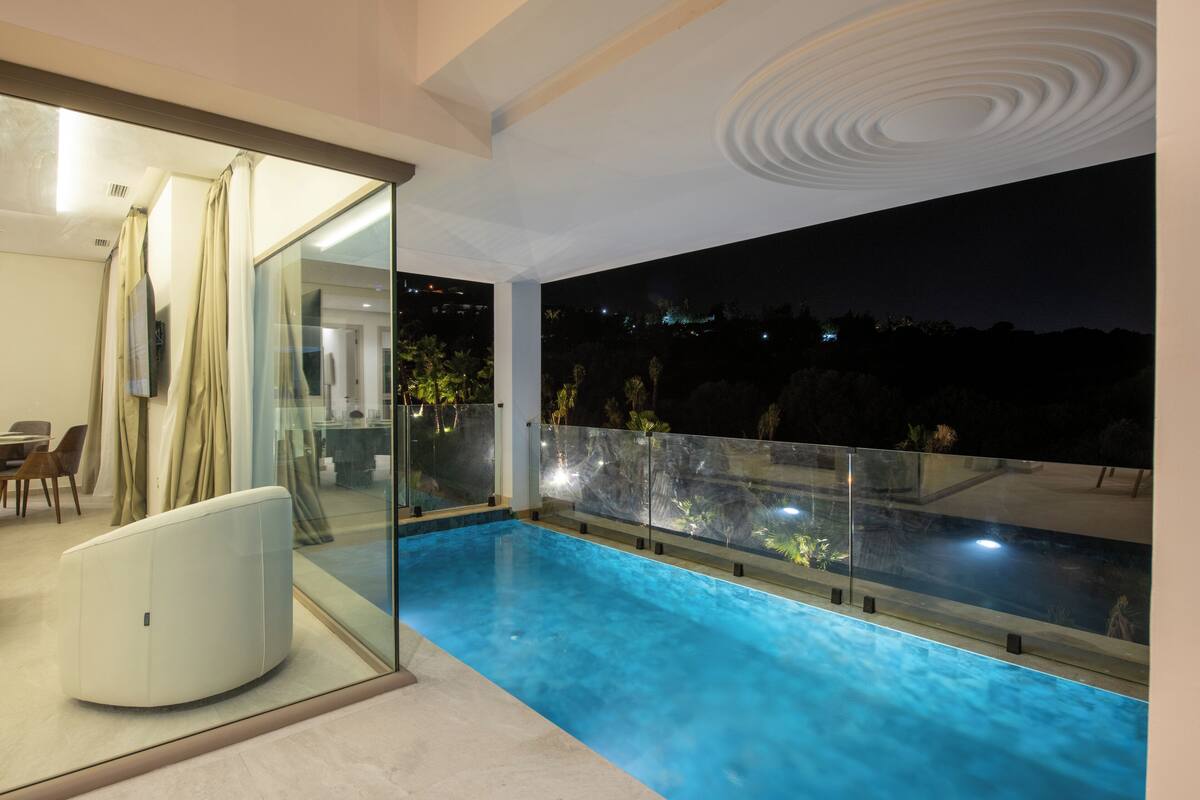 A modern indoor swimming pool with glass walls, a seating area, and a view of the night sky.