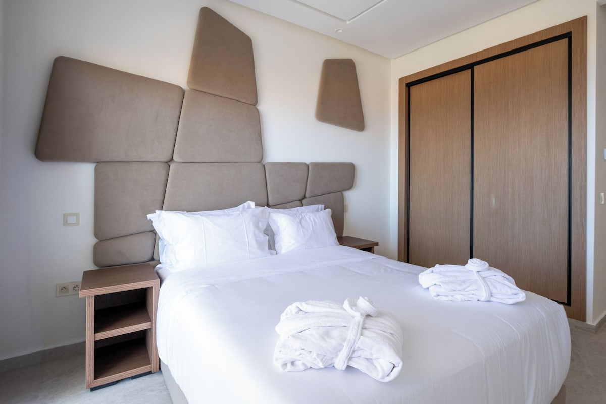Quarto de hotel moderno com uma cama grande, mesa de cabeceira de madeira e um guarda-roupa.