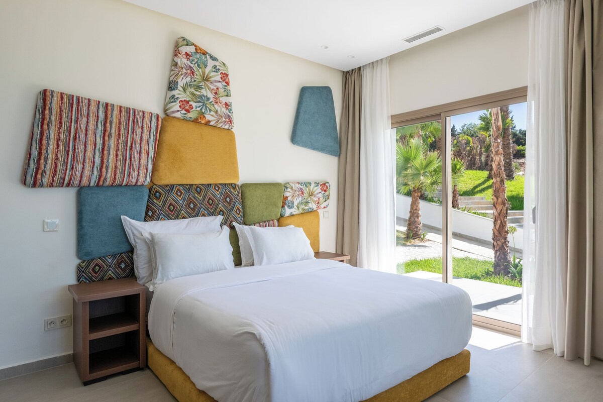 Quarto de hotel moderno com uma cama grande, uma mesa de cabeceira e vista para um jardim através de uma porta de vidro deslizante.