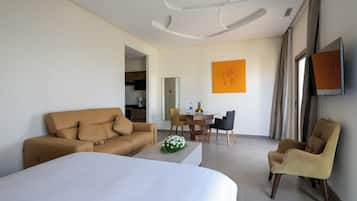 Una habitación de hotel moderna con una cama grande, un sofá color arena, una zona de comedor con mesa y sillas, y un televisor montado en la pared.