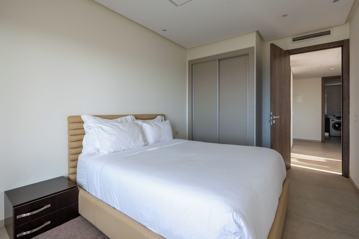 A modern bedroom with a large bed, a bedside table, a wardrobe, and a view into another room.