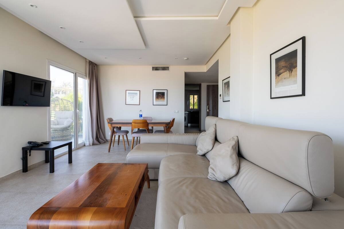 A modern living room with a large beige sofa, a wooden coffee table, a TV mounted on the wall, and a dining area with a table and chairs.