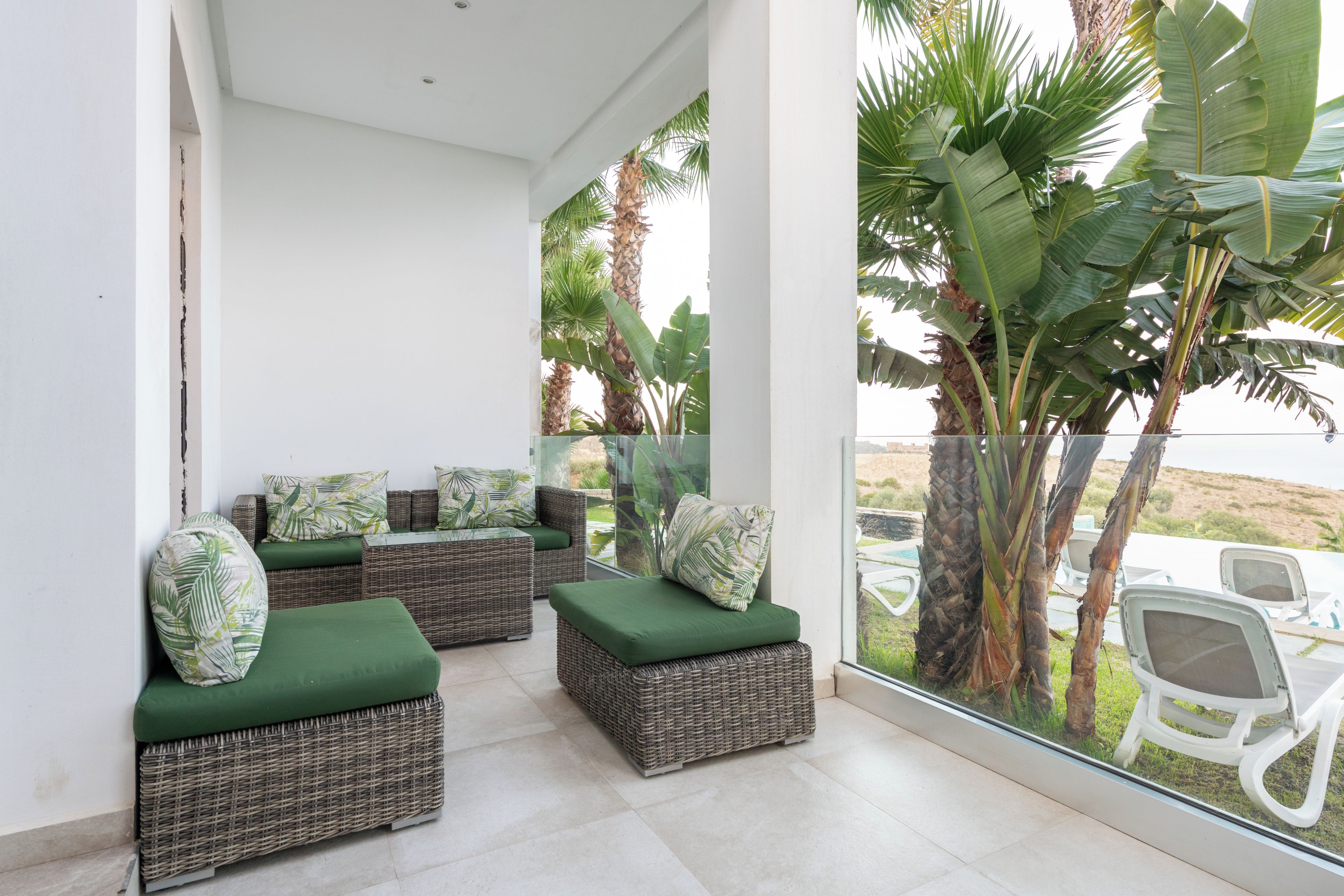 Un balcone con mobili in vimini, cuscini verdi e vista su palme.
