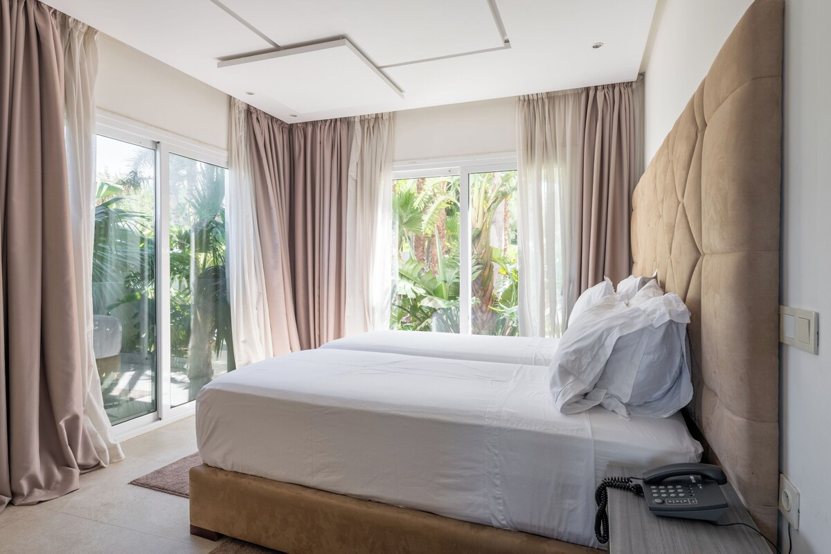 A modern hotel room with a large bed, a bedside phone, and a view of lush greenery through the windows.