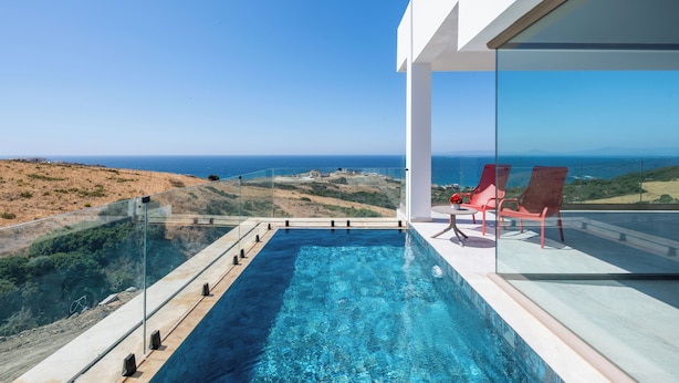 A modern infinity pool with clear blue water, red lounge chairs, and a glass railing overlooking a coastal landscape. - Le Tanger By Mnar Palm (Al Bahraoyine)