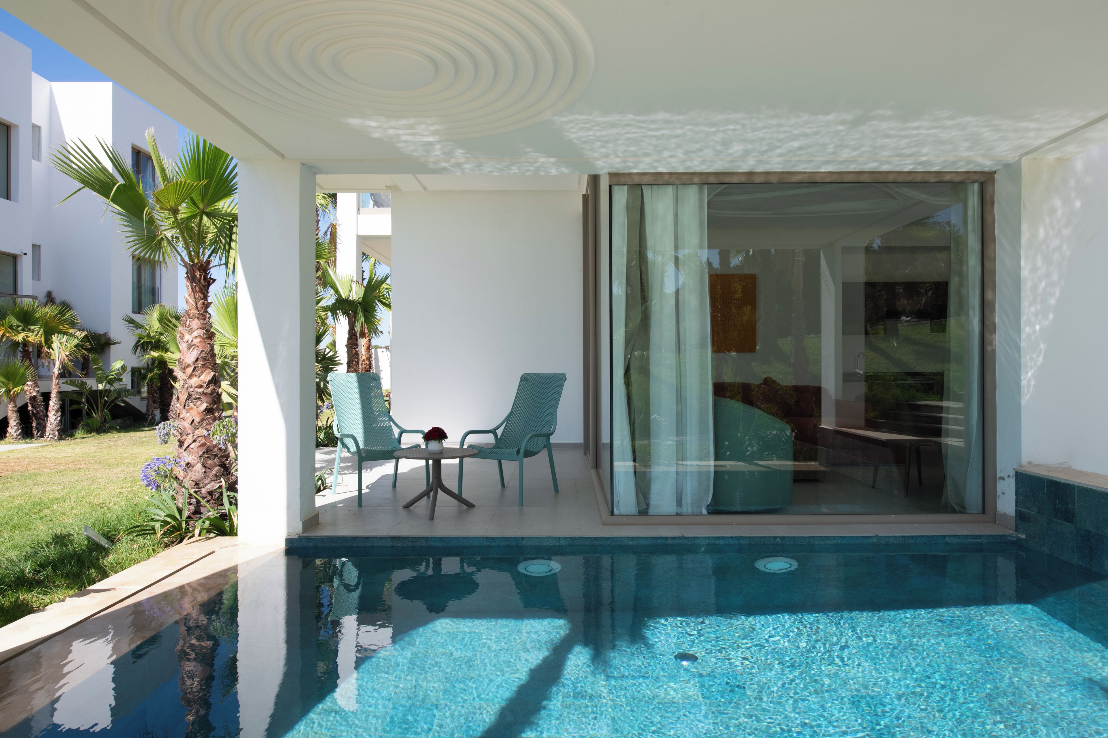 Un área de piscina moderna con agua azul cristalina, una puerta de vidrio que da a una habitación y una pequeña mesa con sillas.