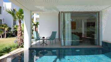 Un área de piscina moderna con agua azul cristalina, una puerta de vidrio que da a una habitación y una pequeña mesa con sillas.