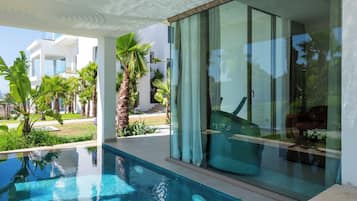 Un área de piscina moderna con agua azul cristalina, rodeada de exuberante vegetación y una puerta de vidrio que da a un espacio interior bien iluminado.