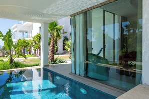 A modern pool area with clear blue water, surrounded by lush greenery and a glass door leading to a well-lit interior space. - Le Tanger By Mnar Palm (Al Bahraoyine)