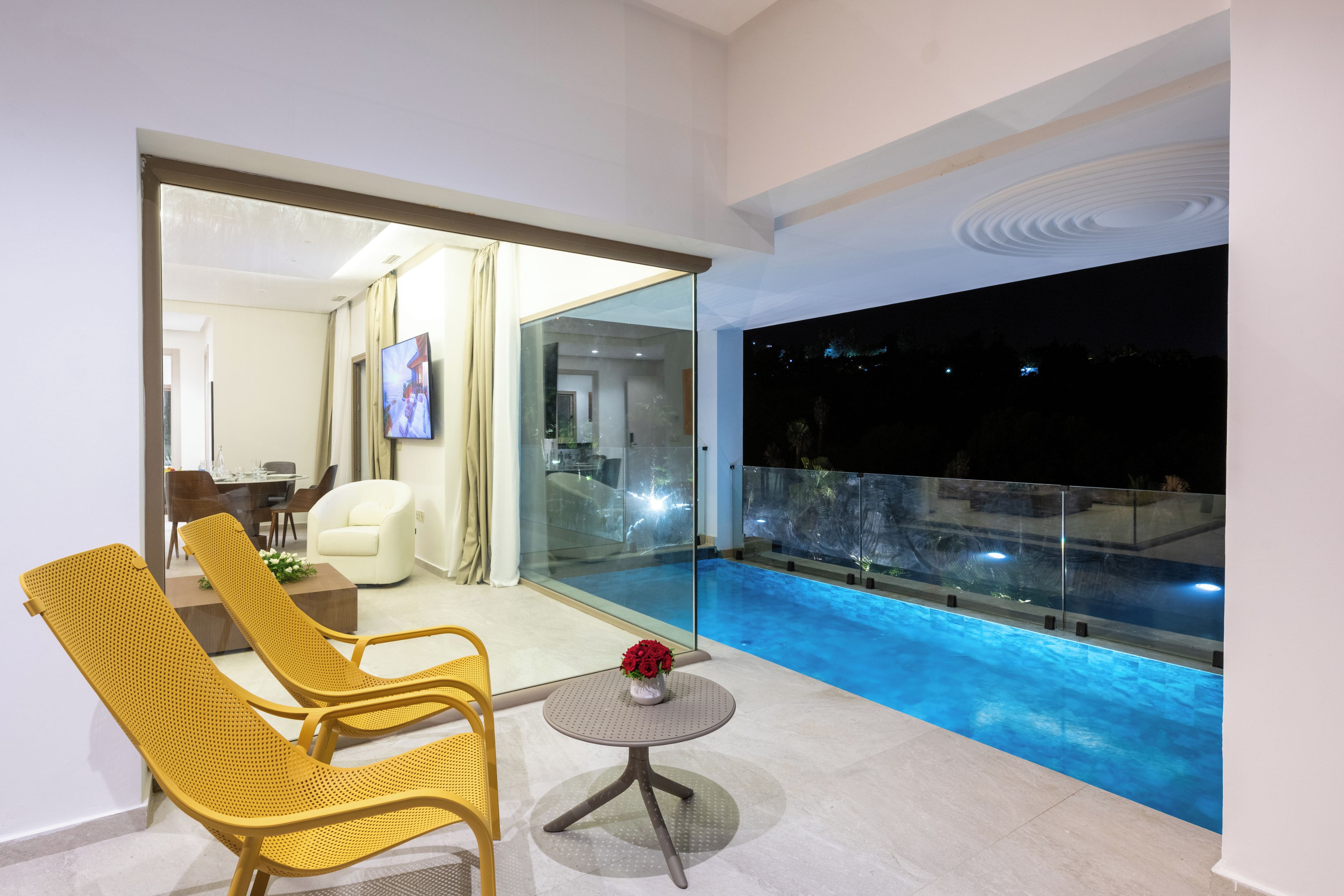 Un balcón moderno con barandilla de vidrio que da a una piscina, una zona de estar con sillas y una mesita, y vistas al interior del edificio.