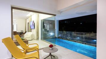 Un balcón moderno con barandilla de vidrio que da a una piscina, una zona de estar con sillas y una mesita, y vistas al interior del edificio.