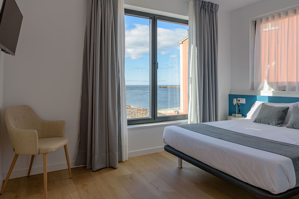 A bedroom with a bed, a chair, a window with a view of the sea, and a nightstand with a lamp.