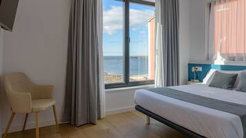 A bedroom with a bed, a chair, a window with a view of the sea, and a nightstand with a lamp.