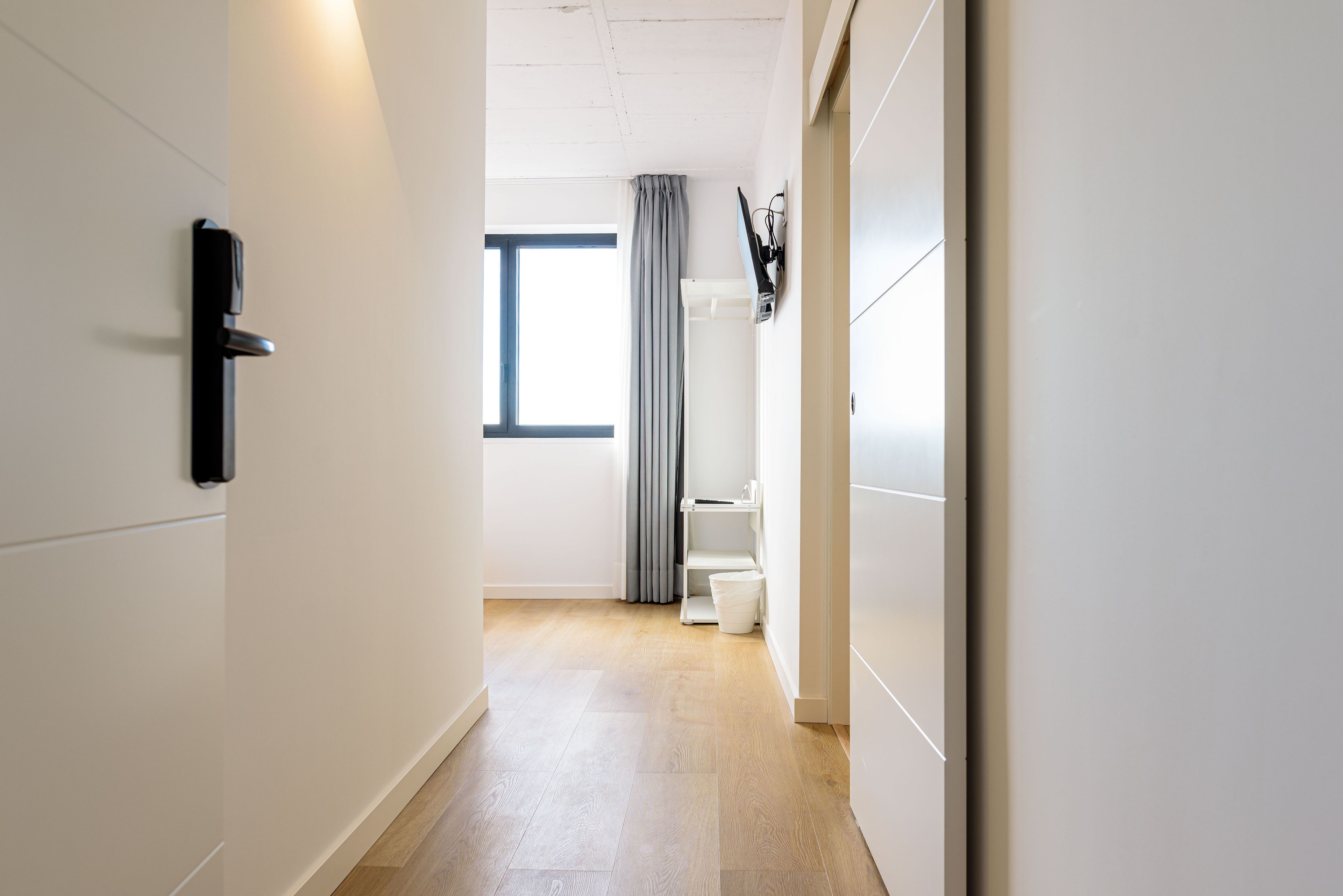 Un pasillo de diseño moderno, con suelo de tonos claros, una puerta con pomo negro y una ventana con cortina gris.