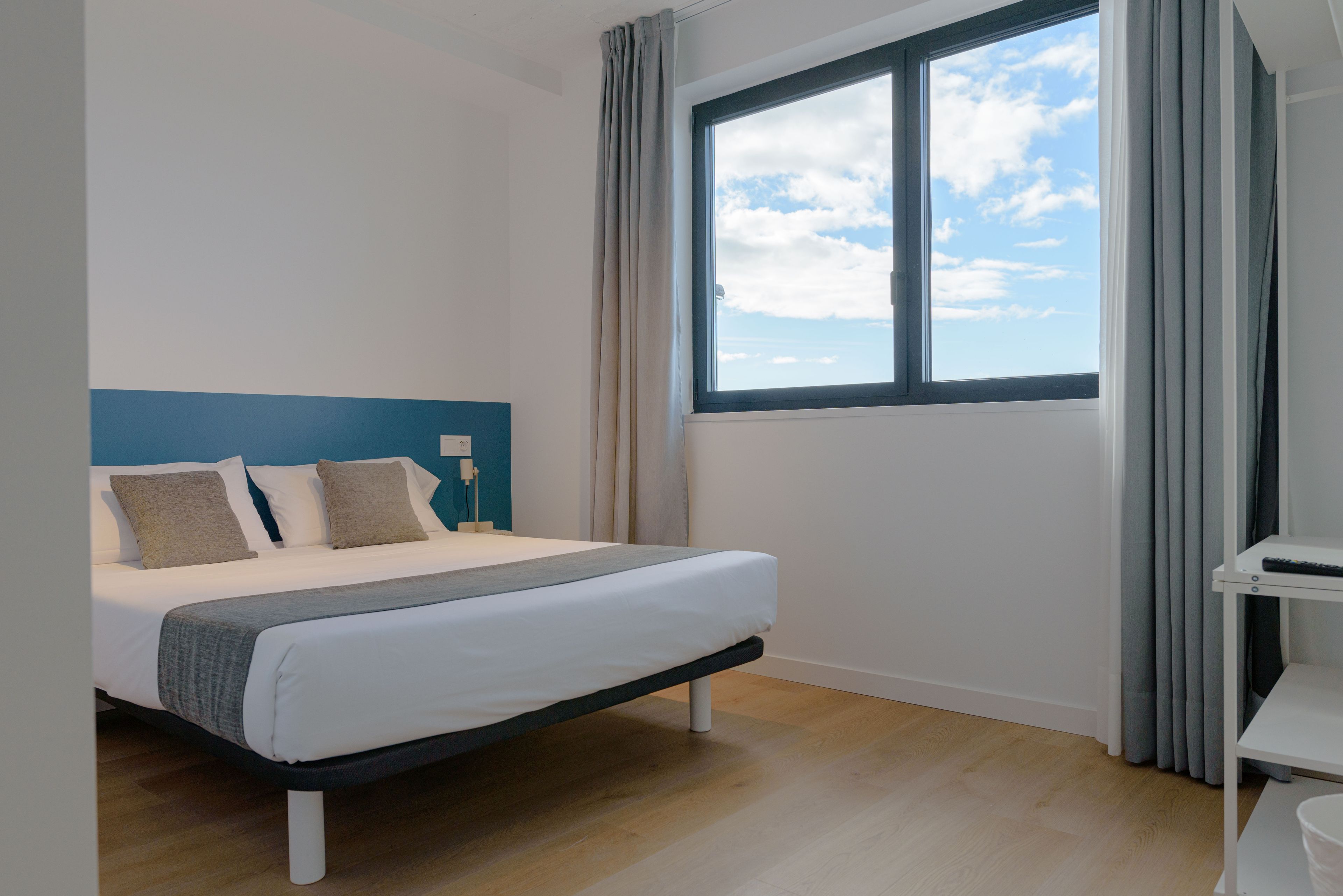 A modern bedroom with a large window, a bed with white bedding, and a blue accent wall.