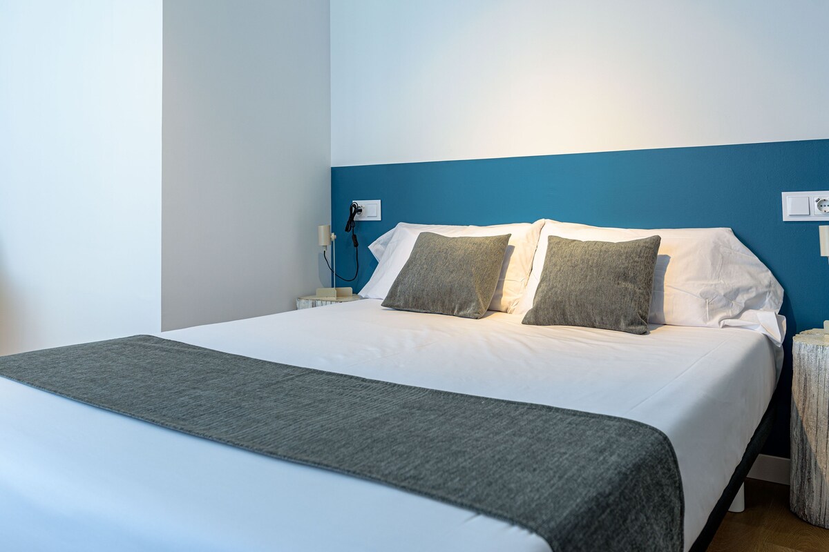 Una cama bien hecha con sábanas blancas y dos almohadas grises, una manta gris y una pared de acento azul.