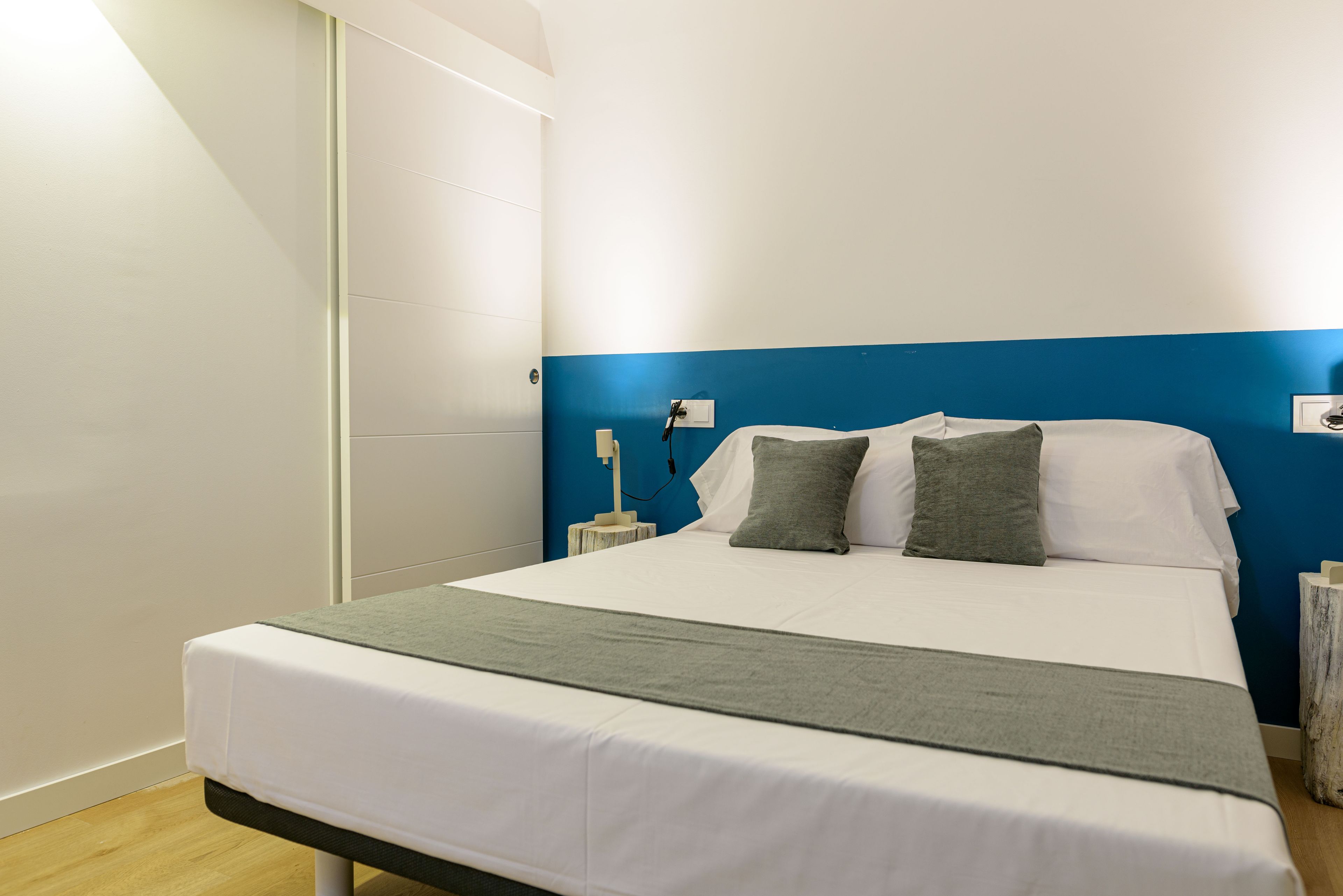 A neatly made bed with white and gray bedding, a blue accent wall, and a bedside table with a lamp.
