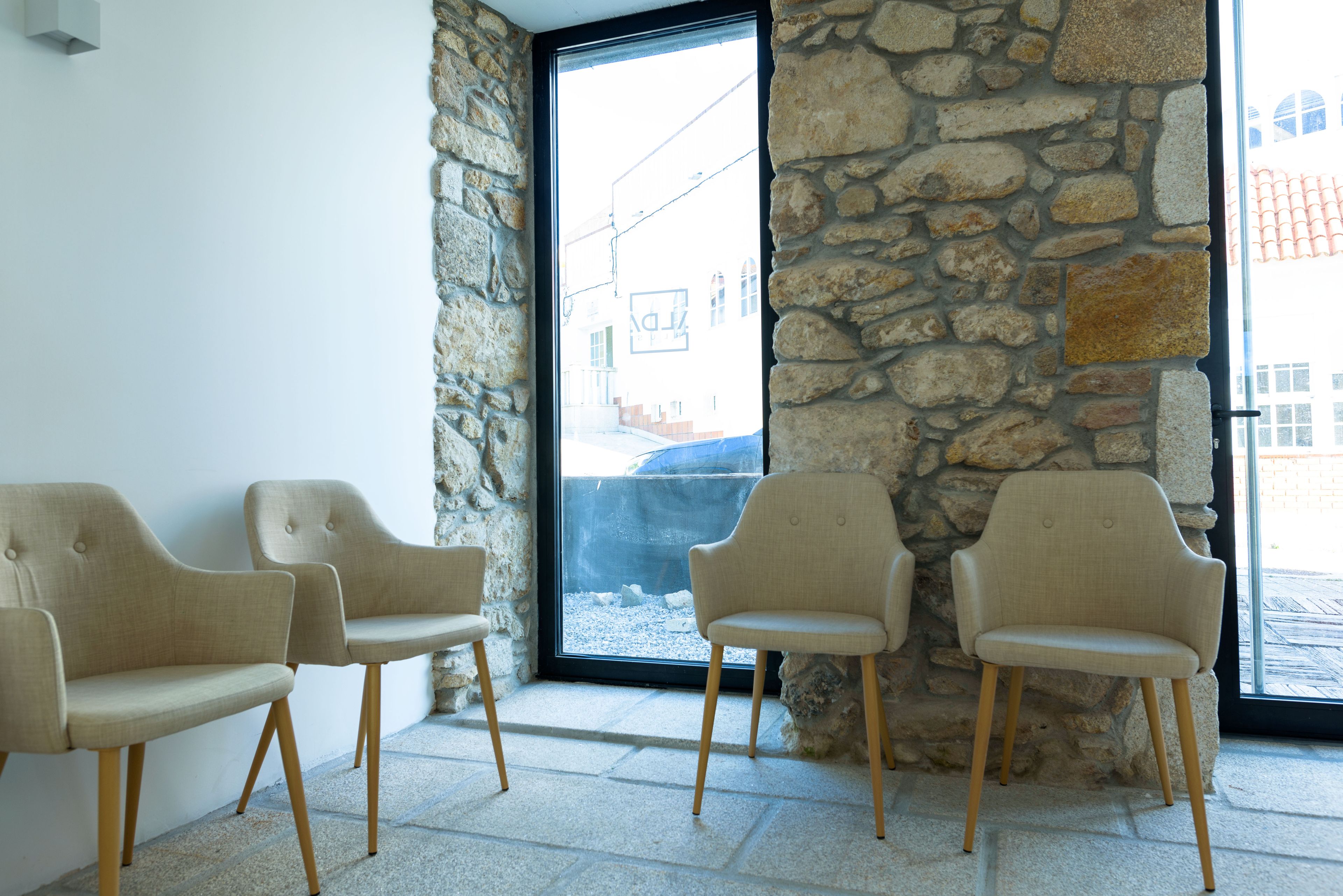 Une pièce avec trois chaises beiges, un mur en pierre et une grande fenêtre donnant sur les bâtiments environnants.