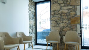 A room with three beige chairs, a stone wall, and a large window with a view of buildings outside. - Alda Corrubedo (Ribeira)
