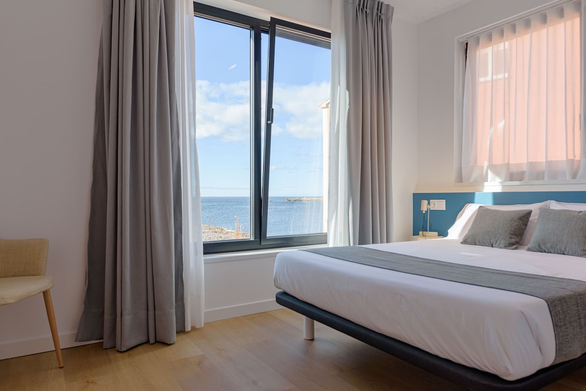 A bedroom with a bed, a chair, a window with curtains, and a view of the sea.