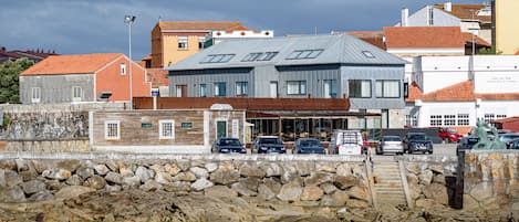 A coastal building with a stone wall in the foreground, a parking area with several cars, and a clear sky.