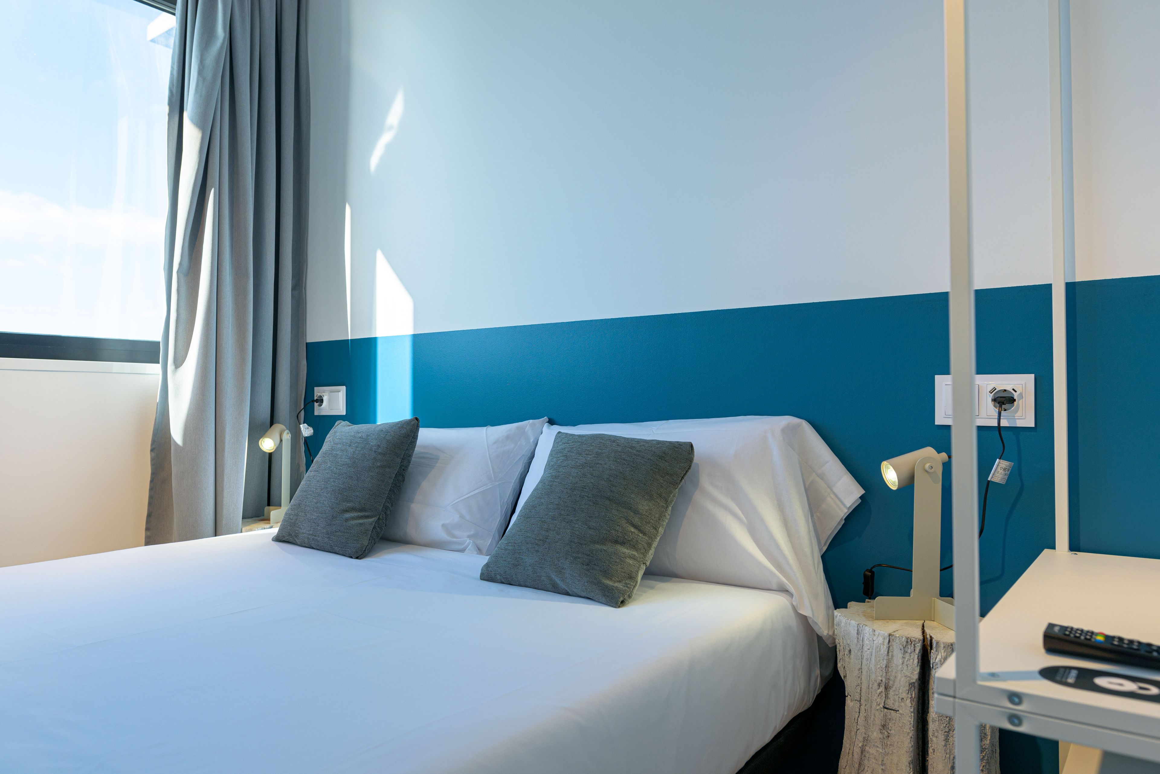Een opgemaakt bed met witte en grijze linnens, een bedlampje en een afstandsbediening op een klein tafeltje naast het bed.