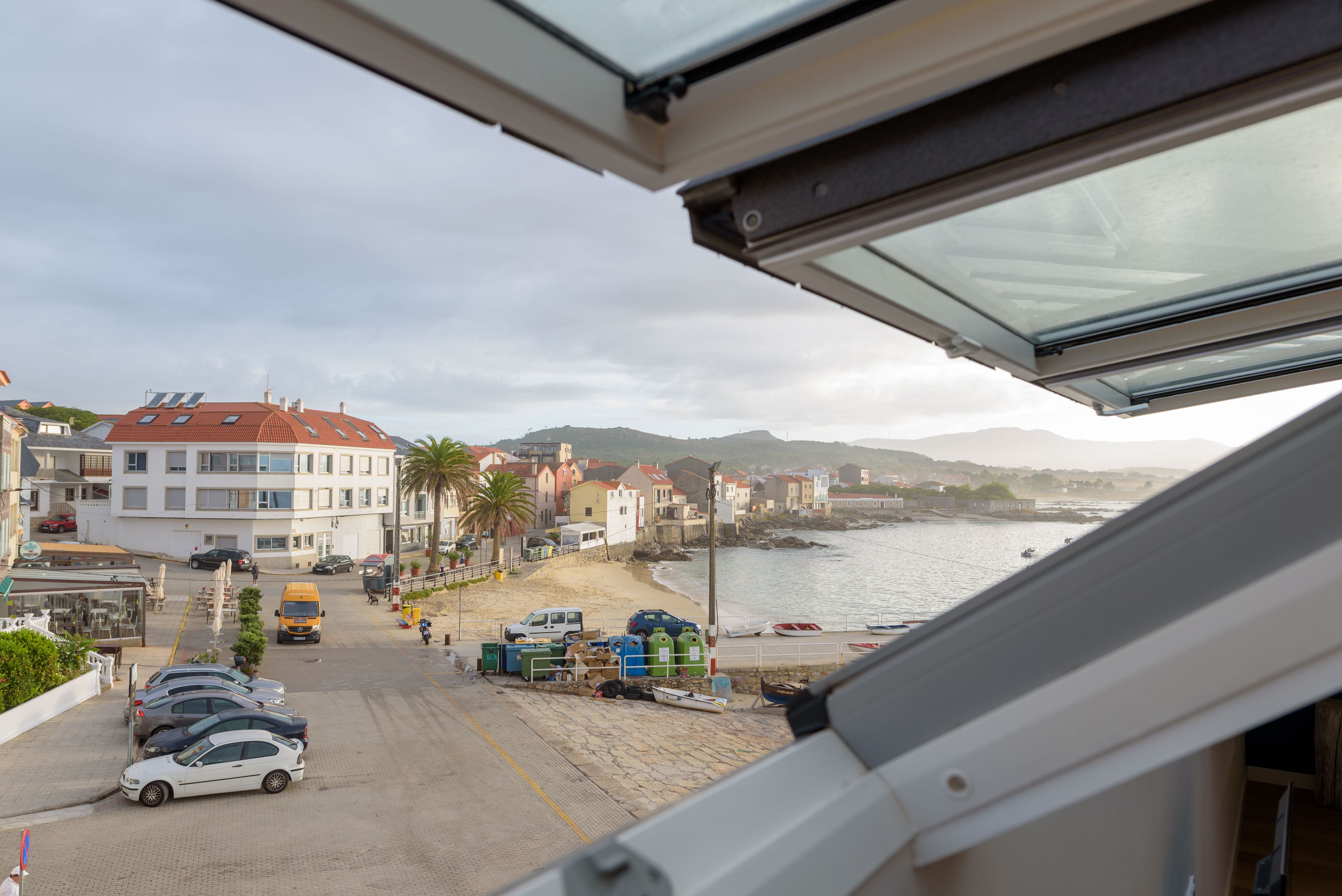 Une vue sur la côte, prise depuis une fenêtre donnant sur une plage où sont garées des voitures, une rangée de bâtiments et une étendue d’eau.
