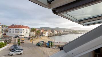 Une vue sur la côte, prise depuis une fenêtre donnant sur une plage où sont garées des voitures, une rangée de bâtiments et une étendue d’eau.