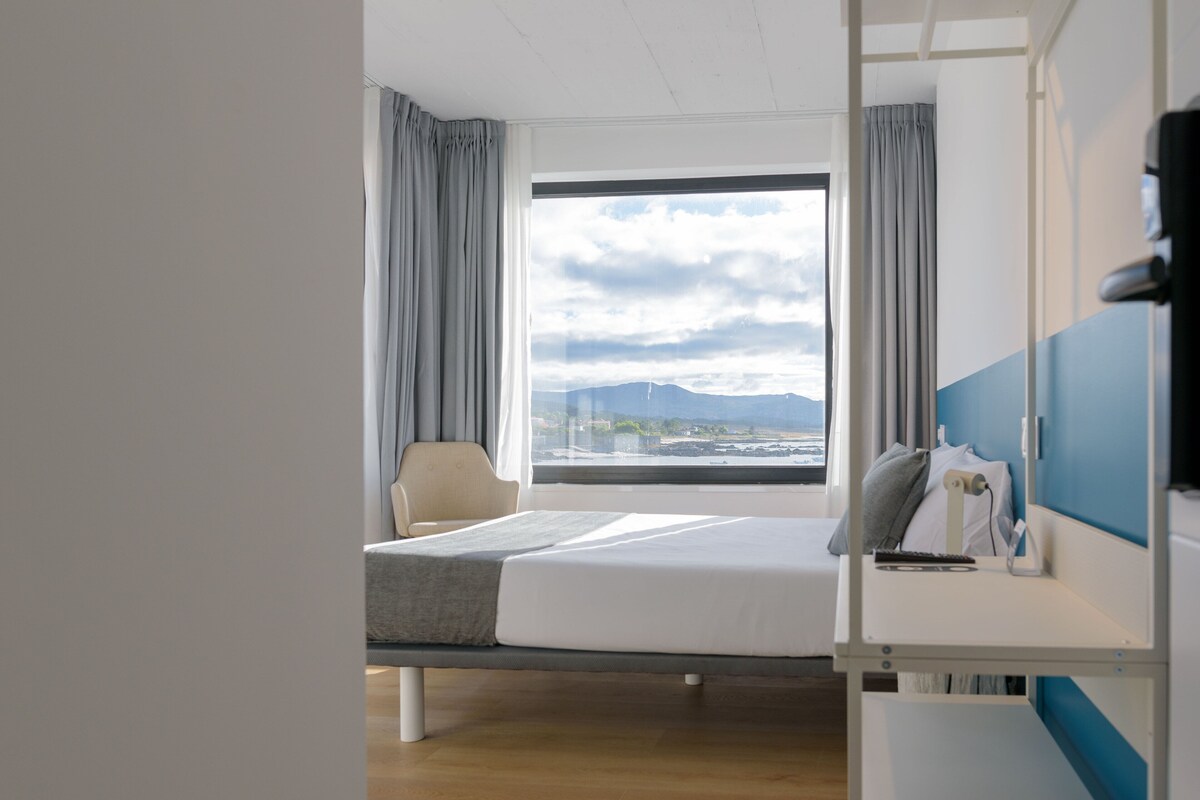 Une chambre d’hôtel équipée d’un lit, d’une chaise, d’un bureau et d’une grande fenêtre donnant sur les montagnes et le paysage urbain.