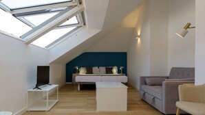 A modern loft bedroom with a sloped ceiling, skylight, a bed with pillows, a grey sofa, a white coffee table, and a TV on a stand. - Alda Corrubedo (Ribeira)