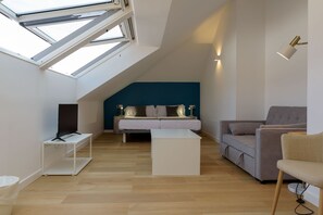 Un dormitorio tipo loft moderno con techo inclinado, tragaluces, una cama con almohadas, un sofá gris, una mesa de centro blanca y un televisor sobre un mueble.