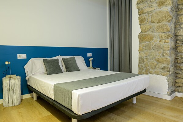 A bed with white bedding, grey pillows, and a grey throw blanket. There is a wooden nightstand with a lamp on it. The room has blue walls, a stone accent wall, and a wooden floor. - Alda Corrubedo (Ribeira)