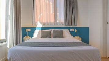 Una cama bien hecha con sábanas blancas y dos almohadas, una mesita de noche con lámpara y una ventana con cortinas.