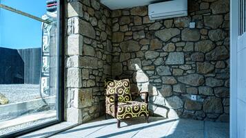 Una habitación con paredes de piedra, un sillón estampado, una ventana y un equipo de aire acondicionado.
