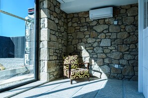 A stone-walled room with a patterned armchair, a window, and an air conditioning unit. - Alda Corrubedo (Ribeira)