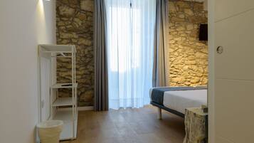 Une chambre avec un mur en pierre, un lit, une étagère blanche et une fenêtre avec des rideaux.