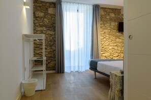 A bedroom with a stone wall, a bed, a white shelf unit, and a window with curtains. - Alda Corrubedo (Ribeira)