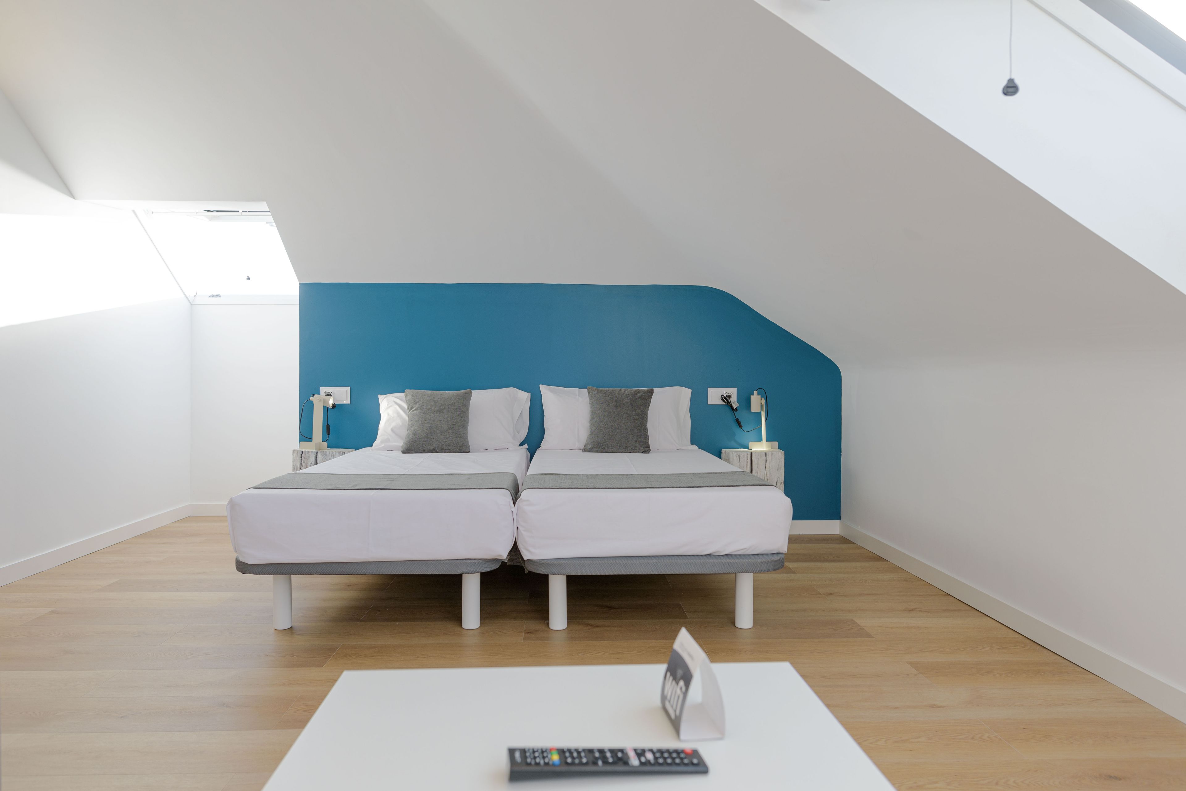 Une chambre moderne avec un plafond en pente, deux lits avec une literie blanche et des oreillers gris, une table blanche avec une télécommande, et un mur d’accent bleu.