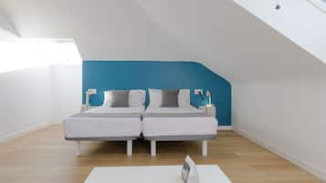 Una habitación moderna con techo inclinado, dos camas con ropa blanca y almohadas grises, una mesa blanca con control remoto, y una pared de acento azul.