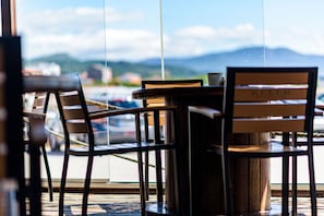 A view through a window with a table and chairs, a rope barrier, and a glimpse of a mountain range in the background. - Alda Corrubedo (Ribeira)