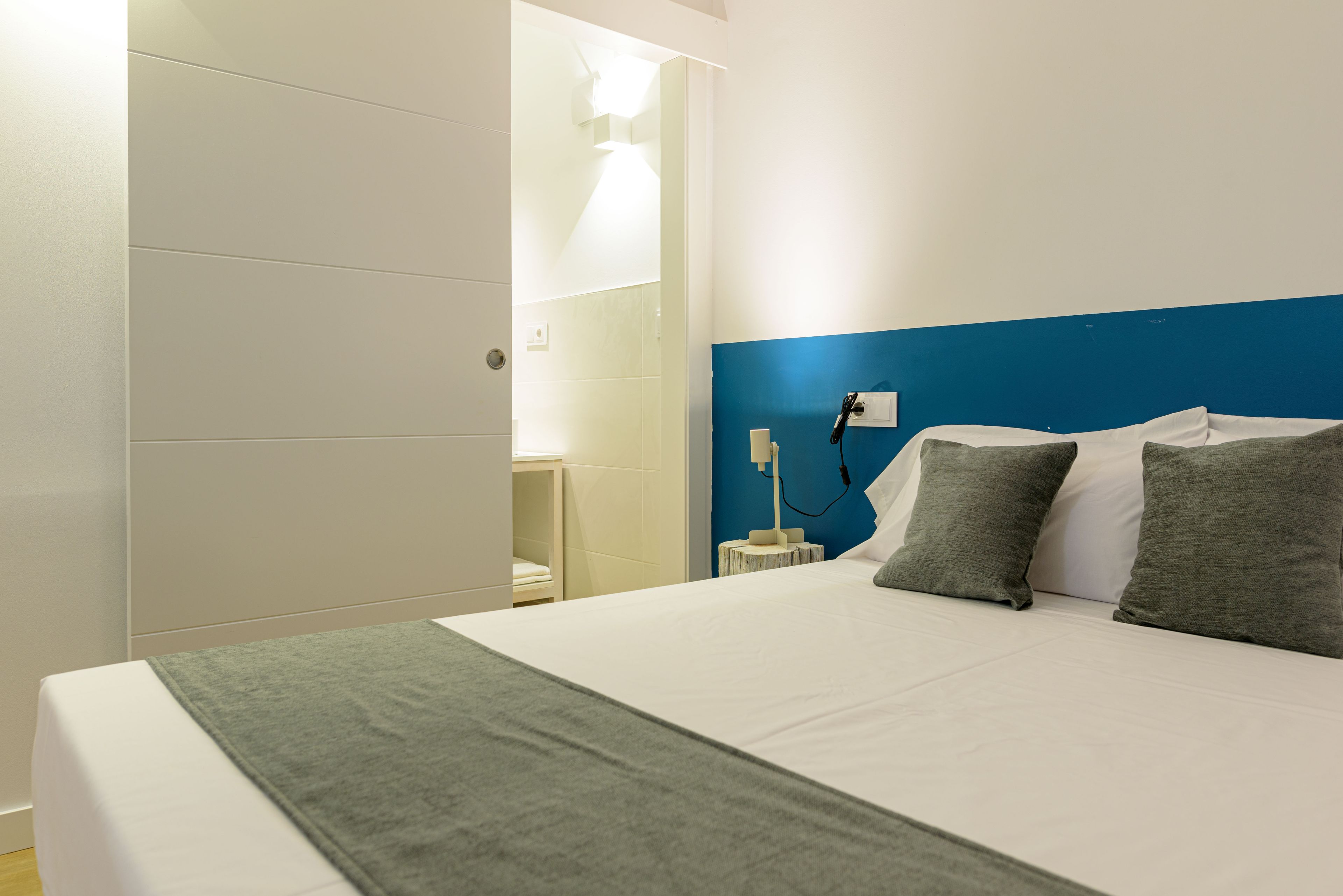 A hotel room with a bed, a nightstand, a lamp, and a bathroom visible through an open door.