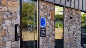 A stone building with a parking sign, a security intercom, and a glass door reflecting greenery. - Alda Corrubedo (Ribeira)