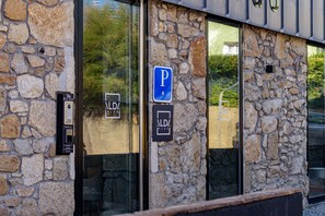 Un edificio de piedra con un cartel de estacionamiento, un interfono de seguridad y una puerta de cristal que refleja vegetación.