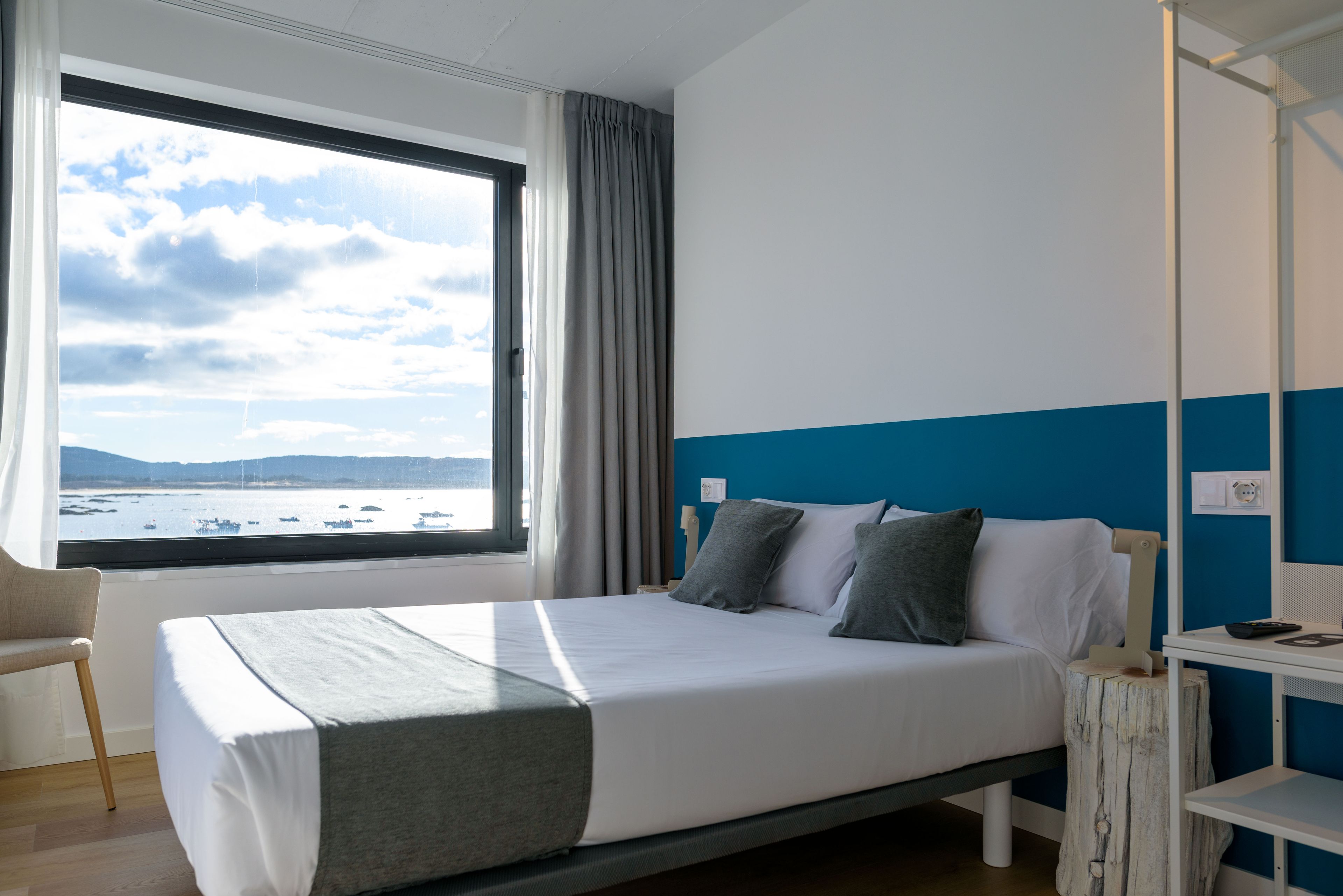 Une chambre d’hôtel avec un lit, une chaise, une table de chevet et une vue sur la mer, visible à travers une grande fenêtre.